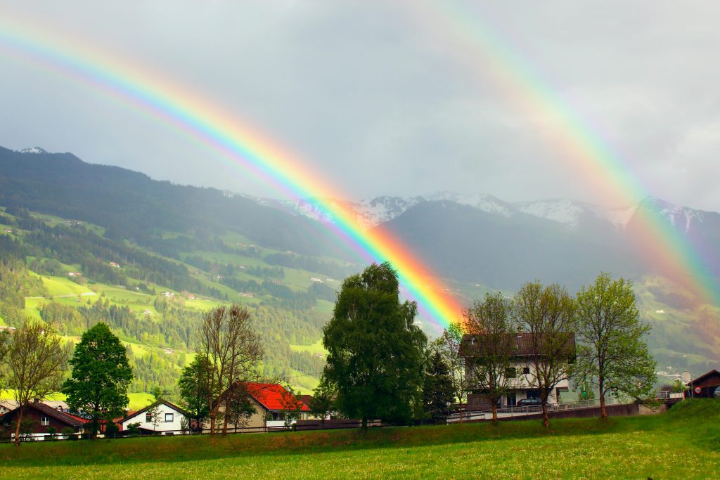 A vibrant rainbow arches over the picturesque village of Schlitters in Tirol, Austria, framed by lush greenery and mountains.